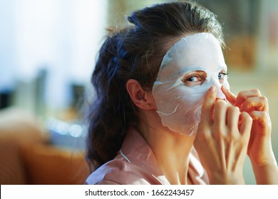 young woman in pajamas at modern home in sunny winter day applying white sheet facial mask.