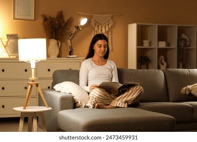 Young woman in pajamas reading magazine on soft couch in living room at night - Powered by Shutterstock