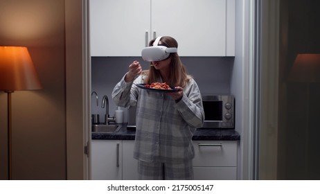 Young woman in pajamas and vr goggles eat pasta standing in kitchen. Female wearing augmented reality goggles eat dinner in virtual restaurant at home - Powered by Shutterstock