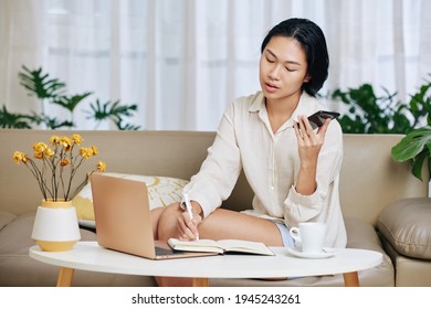 Young Woman In Pajamas Working From Home, She Is Listening To Voice Message And Taking Notes In Planner