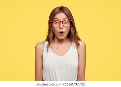 Young woman in panic looks with surprised expression, feels nervous in stressful situation, wears white oversized t shirt, red transparent glasses, opens mouth with stupefaction, isolated on yellow