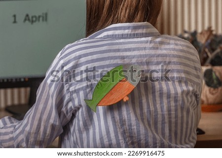 A young woman with a paper fish from behind indoors works at a computer on which the inscription the first of April, April Fool's Day, jokes and jokes in the office on top of each other