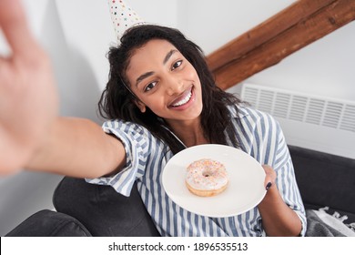 Young Woman In Party Cap Holding Cake And Making Selfie At The Birthday Party Alone At The Bedroom. Girl Smiling To The Camera While Preparing To Blowing Candle At The Doughnut. Stay Home, Quarantine