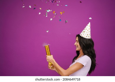 Young Woman In Party Cone Laughing While Blowing Up Party Popper Isolated Over Purple Wall