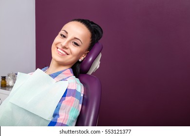 Young Woman Patient In Checkered Shirt With Perfect Straight White Teeth Waiting For Dentist In Dental Chair And Smiling Relaxed, Ready For A Check-up. Beautiful Woman Smile