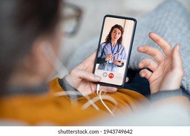 Young Woman Patient In Conversation With Specialist Over Video Conferencing Using Smart Phone. Sick Woman Patient Lying On Sofa Doing Video Consultation With Doctor And Explain Her Symptoms.