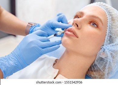 Young woman patient wearing hair cap at beauty clinic cosmetology service close-up lying on medical bed relaxed while doctor doing injections of hyaluronic acid into lips.