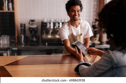 Young Woman Paying By Credit Card At Cafe. Woman Entering Security Pin In Credit Card Reader With Male Barista Standing Behind Checkout Counter At Coffee Shop.