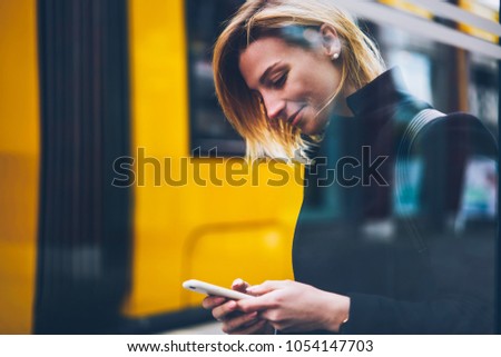 Young woman paying for transport via banking application on telephone waiting for bus, smiling female traveler checking traffic ways and tables on website browsed on smartphone on city street