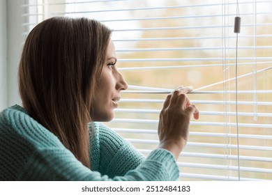 Young Woman peering through blinds sitting indoors on a sunny day, lost in thought and reflection on her surroundings. - Powered by Shutterstock