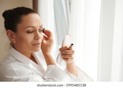 Young Woman With Perfect Make-up Applying Mascara At Her Lashes, Looking At The Small Cosmetic Mirror While Standing In Front Of Window