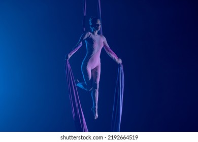 A young woman performing in a circus on aerial silk in the dark with blue light. A female equilibrist balancing on a height. Performance of aerial gymnast.