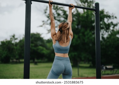 A young woman performs pull ups on outdoor fitness equipment in a park, focusing on her biceps, shoulders, and lateral muscles. - Powered by Shutterstock