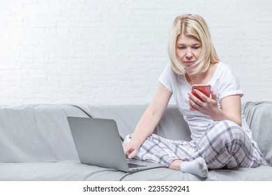 A young woman with a phone and a laptop is sitting on a gray sofa against a white brick wall. Pretty serious blonde in home clothes. Education and work online.