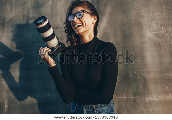 Young woman photographer with her professional\
camera smiling against oliphant backdrops. Woman photographer with\
digital camera.