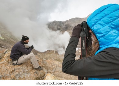 Young Woman Photographs Man In Crater Active Volcano Surrounded By Smoke Hot Springs, Eruption Fumaroles, Gas-steam Volcanic Activity. Kamchatka Peninsula, Russian Far East - September 8, 2014