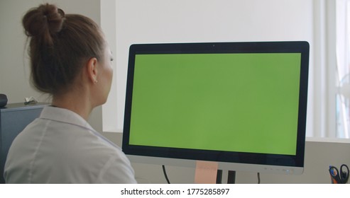 Young woman physician work on computer with green mockup screen. Girl in uniform and lab coat clothes sitting in hospital office room using laptop. Over shoulder camera shot