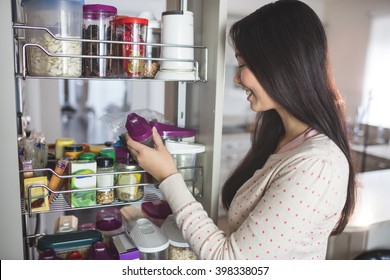 Young Woman Picking A Bottle From Storage Cabinet In Kitchen