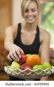 A Young Woman Is Picking Up A Piece Of Fruit, Smiling, And Looking Away From The Camera.  Vertically Framed Shot.