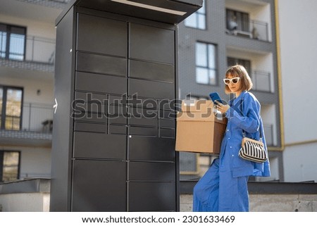Young woman picks up parcels from automatic post office machine, standing with phone near apartment building. Concept of fast delivery to automatic self lockers