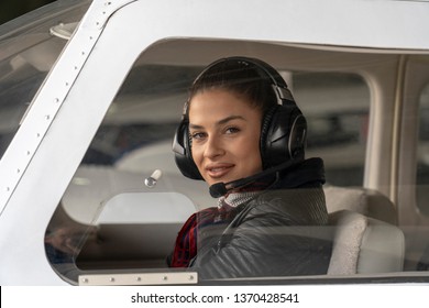 Young Woman Pilot With Headset Looking Through The Cockpit Window. Portrait of attractive young woman pilot with headset. She is looking at camera through  the cockpit window of a private plane.