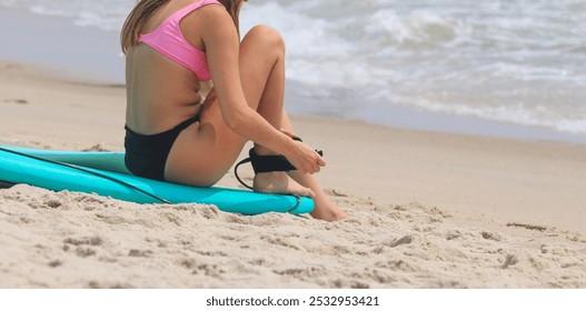 A young woman in a pink bikini prepares for a surfing session by securing her surfboard leash on the beach. - Powered by Shutterstock