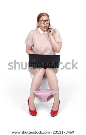 Young woman in a pink dress and glasses is talking on a smartphone sitting on the toilet, a laptop on her lap, isolated on a white background