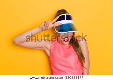Young woman in pink dress and sun visor posing against yellow background and showing peace sign.