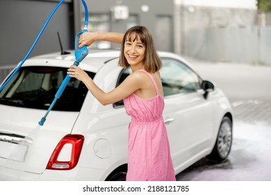 Young Woman In Pink Dress Washing Her Tiny Vehicle With Washing Gun Under The Pressure At Self-service Car Wash. Easy Process Of Car Care By Female Concept