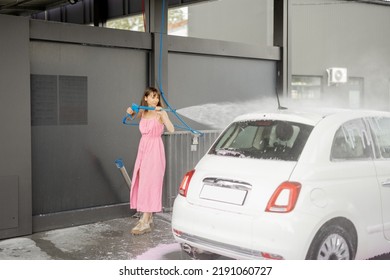 Young Woman In Pink Dress Washing Her Tiny Vehicle With Washing Gun Under The Pressure At Self-service Car Wash. Easy Process Of Car Care By Female Concept