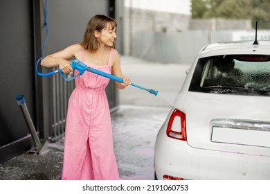 Young Woman In Pink Dress Washing Her Tiny Vehicle With Washing Gun Under The Pressure At Self-service Car Wash. Easy Process Of Car Care By Female Concept