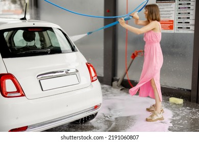 Young Woman In Pink Dress Washing Her Tiny Vehicle With Washing Gun Under The Pressure At Self-service Car Wash. Easy Process Of Car Care By Female Concept