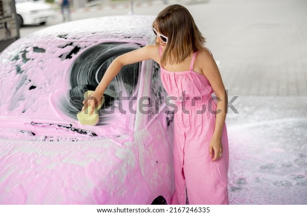 Young woman in pink dress wiping her tiny car\
covered in nano foam with a sponge at car wash. Concept of easy and\
beautiful self-service at car\
wash