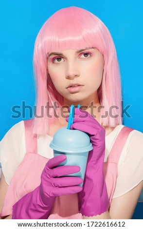 Young woman with pink hair drinking from disposable blue plastic cup. Teenager holding summer cocktail in protective gloves on blue background. Advertising during coronavirus concept