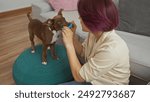 A young woman with pink hair plays with her brown dog indoors, seated on a teal pouf in a cozy living room.