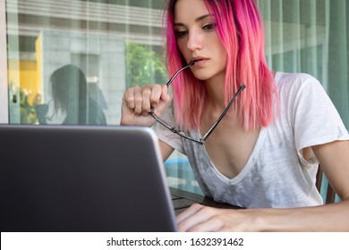 Young woman with pink hair working with laptop in cafe or home terrace. Serious pensive hipster girl freelancer work hard.