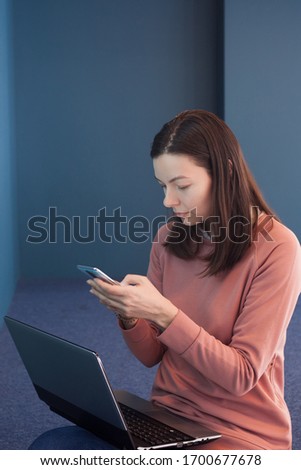 young woman in a pink jumper works in a coworking, blue background. Use develop and configure cross-platform applications. A young female freelancer with a phone in her hands and a laptop on her lap