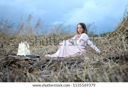 Young woman in pink long dress with lantern sitting on the grey grass. Blue deep sky as a background.
