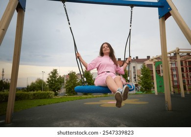 A young woman in a pink outfit swings on a blue tire swing in a city park - Powered by Shutterstock