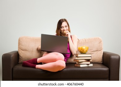 Young Woman In A Pink Outfit, Working On A Laptop While Sitting On The Couch At Home, Eating Potato Chips Placed On The Books. Gray Background, Easy To Remove.