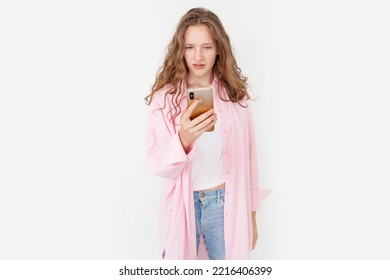 Young Woman In A Pink Shirt Looks Angrily At The Phone Screen. Sad Teen Girl With Mobile Phone Isolated On White Background