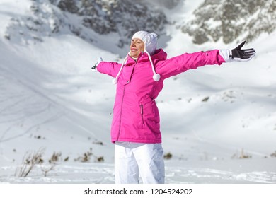 Young Woman In Pink Ski Jacket, Gloves And Pants, Arms Spread, Eyes Closed, Smiling Facing Sun, Snow Covered Mountain Behind Her.