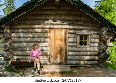 A young woman in a pink sweatshirt and denim shorts sits on a bench attached to a rustic log cabin in a sunny forest setting. This photo captures the essence of outdoor relaxation and rustic charm - Powered by Shutterstock