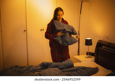 Young woman in plaid pajamas preparing the bed for sleep, fluffing the pillow in a cozy bedroom with warm light." - Powered by Shutterstock