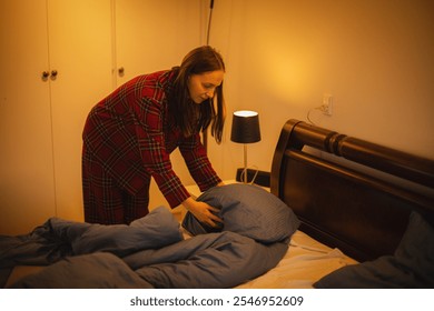 Young woman in plaid pajamas preparing the bed for sleep, fluffing the pillow in a cozy bedroom with warm light." - Powered by Shutterstock