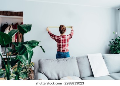 Young woman in plaid shirt doing measuring with a measure tape on the wall. Girl wants to put a picture on the wall at home. Housekeeping work. Doing repair herself. DIY, equality in work concept. - Powered by Shutterstock