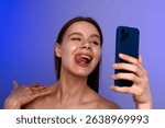 A young woman playfully poses for a selfie with a blue smartphone, sticking her tongue out against a blue background.