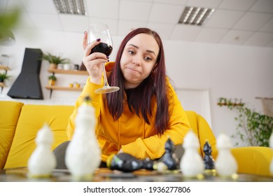 Young Woman Playing Chess And Drinking Red Wine In Room. Adult Female With Alcohol Plays In Logical Board Game With Herself, Sitting On Sofa