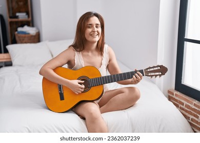 Young woman playing classical guitar sitting on bed at bedroom - Powered by Shutterstock