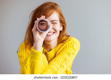 Young woman playing I spy with the camera looking through a rolled cylinder of paper with one eye and a happy smile. Redhead woman looking through paper telescope 